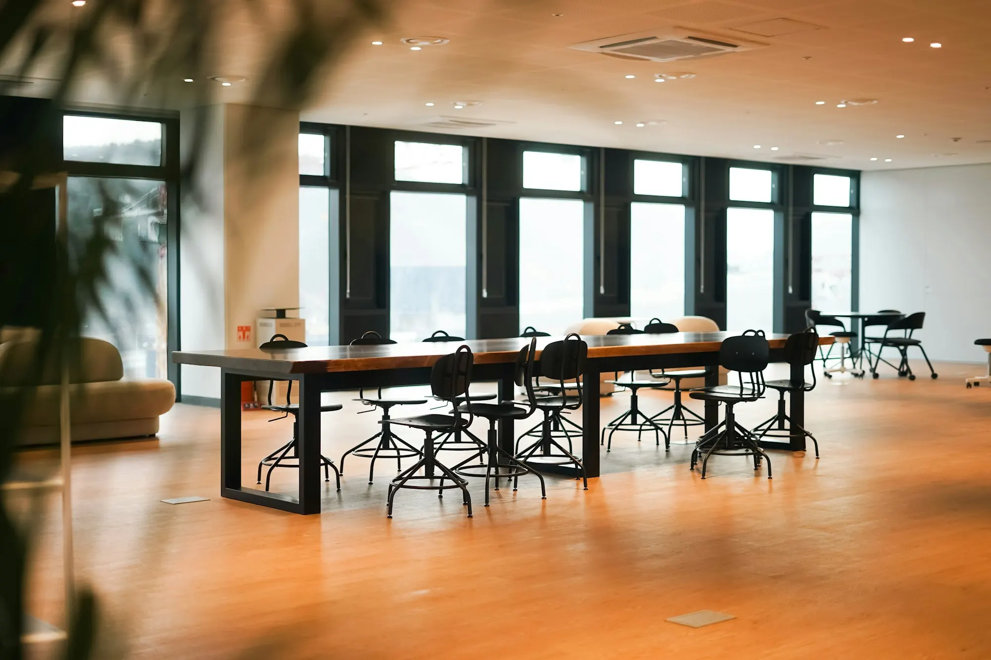 Plateau de bureaux : grandes tables en bois et tabourets noirs sur un sol en bois blond, façade vitrée.