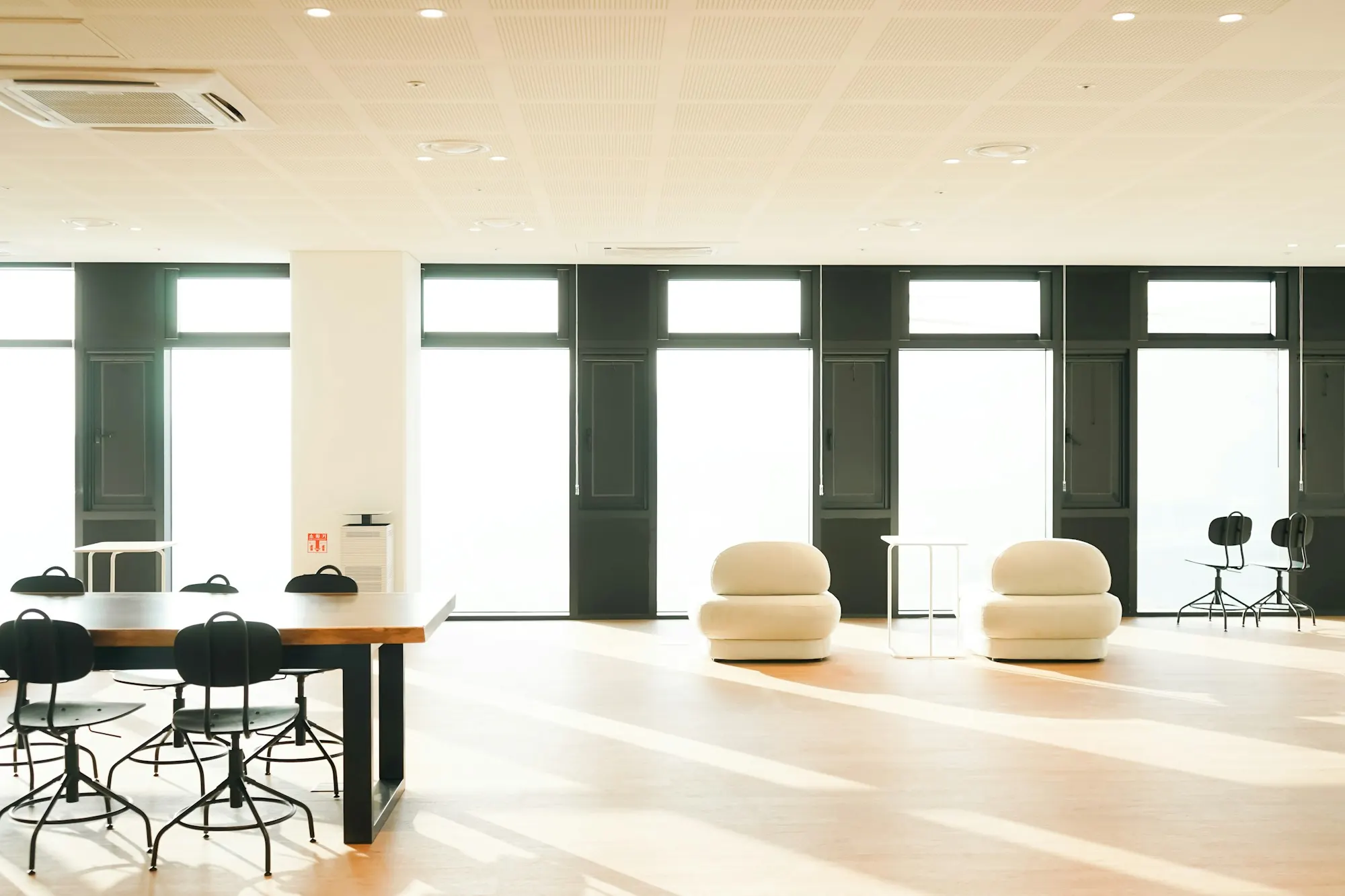 Plateau de bureaux clair sur sol en bois : grandes tables à piètement noir, poufs blancs, façade vitrée.