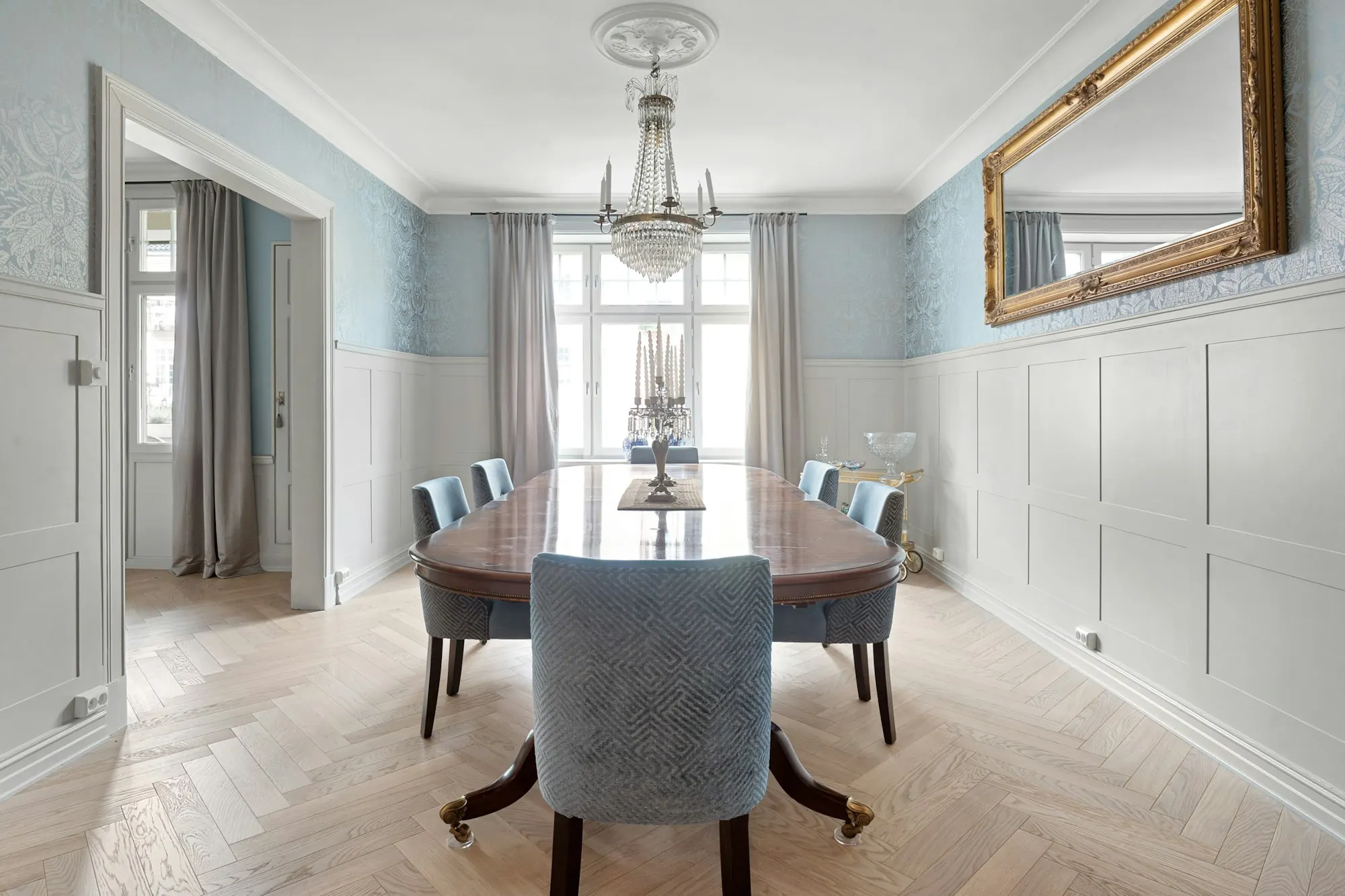 Salle à manger à boiseries blanches : parquet clair à bâtons rompus, table ovale, lustre en cristal et grand miroir doré.