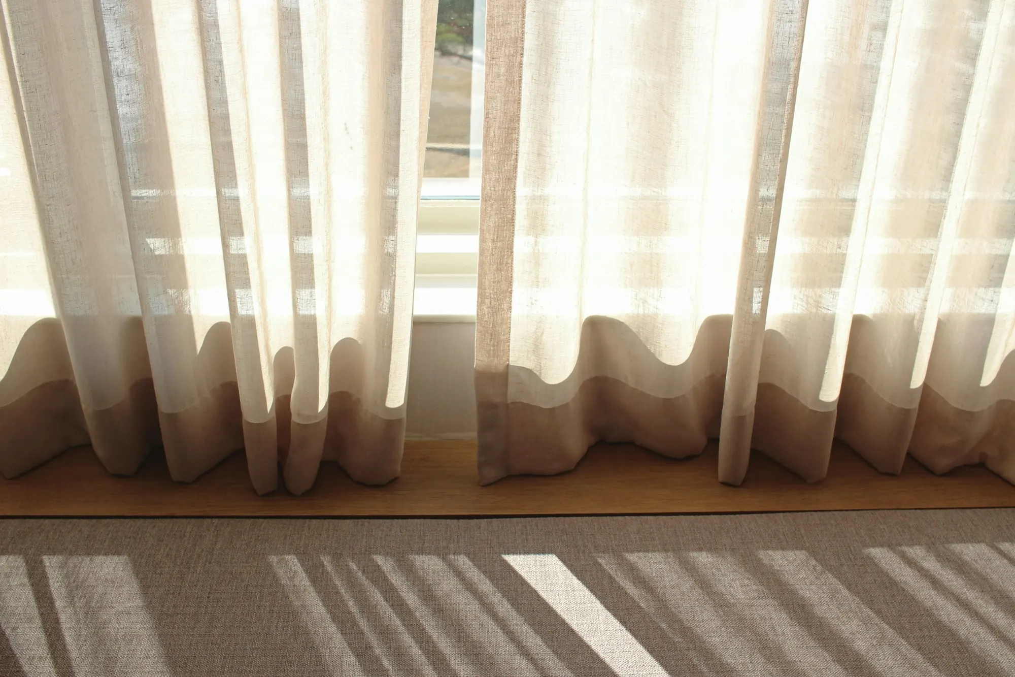 Voilages de lin écru en contre-jour devant une fenêtre, ombres de lattes sur un tapis.