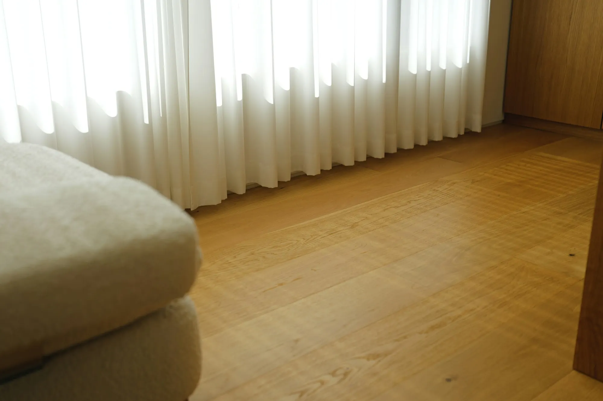 Voilages blancs filtrant la lumière sur un parquet blond à lames larges, accoudoir de canapé au premier plan.